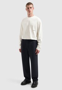 Witte langesleeve hemd met een klein zwart logo op de borst, gecombineerd met donkere, brede pijpen broeken en chunky zwarte schoenen.