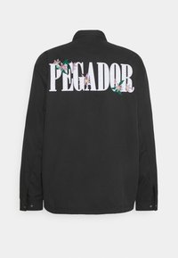Camicia nera con colletto; presenta grande scritta ricamata in bianco "PEGADOR" e accenti floreali sul retro. Maniche con bottoni.