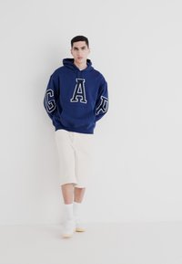Felpa blu navy con lettering bianco "GAP", vestibilità comoda, materiale in cotone. Abbinata a pantaloni corti crema e sneaker bianche. Design pulito.