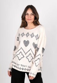 Hellbeige oversized Pullover mit grauen geometrischen und Herzmustern. Hat einen weiten Halsausschnitt und gerippte Bündchen, hergestellt aus weichem, strukturiertem Material.