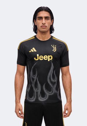 Man draagt een zwart Juventus voetbalshirt met gouden logo’s, strepen op de schouders en vlammenpatronen aan de voorkant tegen een lichte effen achtergrond.