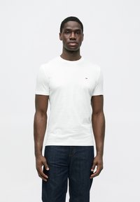 Λευκό βαμβακερό T-shirt με στρογγυλή λαιμόκοψη και κοντά μανίκια, με μικρό λογότυπο στο στήθος. Φορεμένο με σκούρο μπλε τζιν.