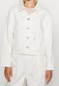 Vitt denimjacka med krage, fyra framfickor och silverfärgade knappar. Synliga sömmar och kortad design.