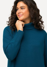 Minkštas tekstūruotas teal spalvos golfas su ilgomis rankovėmis ir raštuotais akcentais. Paprastas, laisvas dizainas, be matomų raštų.