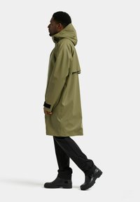 Olivengrøn vandtæt regnfrakke med hætte, der har en løs pasform, sorte detaljer ved manchetterne, og kombineret med sorte bukser og sorte støvler.