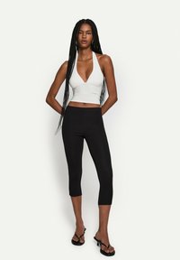 Hvid halterneck crop top med en dyb udskæring, parret med sorte cropped leggings; glat tekstur med en tætsiddende pasform. Sorte flip-flops.