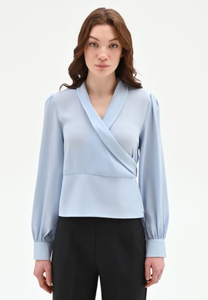 Donna con capelli castano ondulati indossa una blusa avvolgente a maniche lunghe di colore azzurro chiaro e pantaloni neri su sfondo bianco semplice.