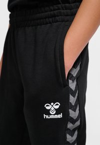 Schwarze Jogginghose mit einem strukturierten Zickzackmuster an den Seiten, versehen mit einem weißen Logo auf der Vorderseite. Elastischer Bund und seitliche Taschen.