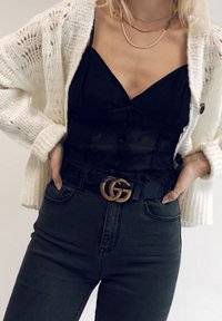 Zwarte blouse met knoopjes en een gerimpelde zoom onder een witte gebreide cardigan. Zwarte jeans, geaccentueerd door een gouden GG-buckle. Eenvoudige gelaagde kettingen.