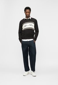 Νέος άνδρας όρθιος με τα χέρια στις τσέπες, φοράει μαύρο πουλόβερ με λευκή επιφάνεια και επιγραφή, λευκό πουκάμισο, σκούρο τζιν και λευκά αθλητικά παπούτσια.