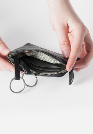 Schwarze Leder Geldbörse mit Metallreißverschluss, ausgestattet mit zwei Schlüsselringen und einem gemusterten Innenfutter. Die Hände halten die Geldbörse offen.