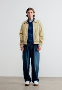 Νεαρός άνδρας που στέκεται φορώντας μπεζ μπουφάν, μπλε ριγέ πουκάμισο, φαρδιά σκούρα τζιν και λευκά αθλητικά παπούτσια μπροστά σε απλή λευκή τοίχο.