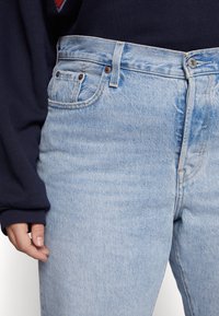 Zbliżenie na osobę w jasnoniebieskich dżinsach i ciemnogranatowej bluzie z długim rękawem, z lewą ręką swobodnie opartą przy boku.