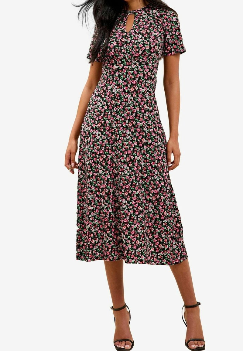 Robe midi fleurie à manches courtes, fond noir, fleurs roses et blanches, encolure ronde avec détail goutte, silhouette évasée fluide.