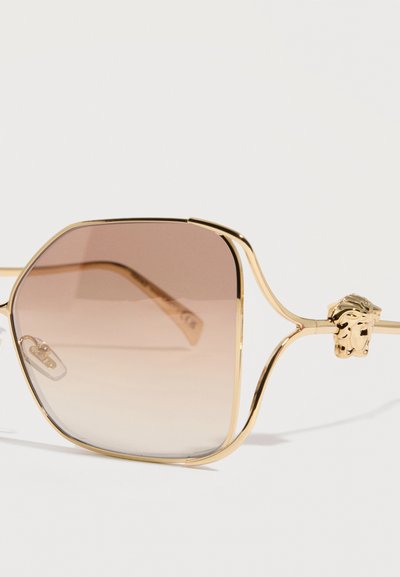 Τετράγωνα φακοί με καφέ βαθμιδωτό χρώμα, λεπτά μεταλλικά χρυσά πλαίσια και διακοσμητική λεπτομέρεια κεφαλής λιονταριού στο μπράτσο του βραχίονα.