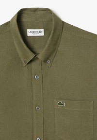 Camicia in lino verde oliva con colletto button-down, tasca frontale e un piccolo logo di coccodrillo ricamato. Presenta bottoni grigi e vestibilità regolare.