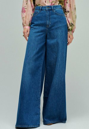 Vaqueros azules de talle alto y pierna ancha, combinados con una blusa transparente de manga larga con estampado floral.