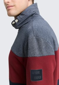 Kraag sweatshirt met een grijze bovenkant en burgundy en marineblauwe horizontale strepen. Inclusief een label op de mouw met merkinformatie.