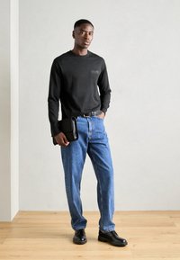 Černá dlouhá rukávová košile s předním logem, modré denimové džíny a černé kožené boty. Modelka drží černou večerní kabelku.