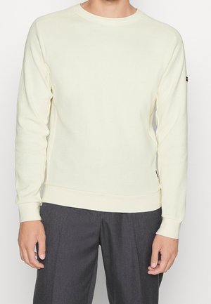 Homme portant un pull crème à col rond et manches longues, ainsi qu'un pantalon gris foncé, debout devant un fond blanc uni.