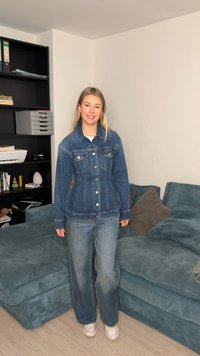 Νεαρή γυναίκα που χαμογελάει, φορώντας ένα τζιν σακάκι και τζιν παντελόνι, στέκεται δίπλα σε έναν μπλε καναπέ από κορδόνι με μαξιλάρια σε ένα καθιστικό.