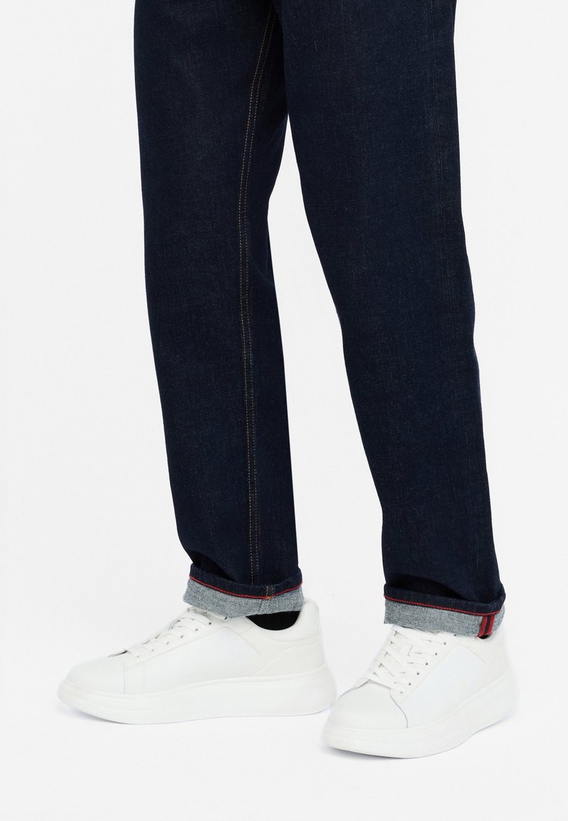Sneakers bianche con una texture di pelle liscia, dotati di sei occhielli e una suola leggermente rialzata, abbinati a jeans di denim scuro con risvolti.