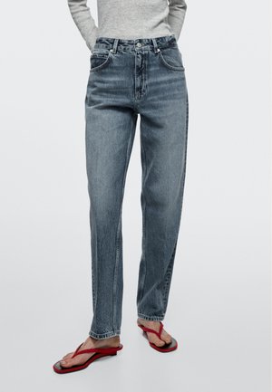 Osoba w szarych, prostych, wysokich dżinsach, włożonym szarym swetrze i czerwonych sandałach typu thong, stojąca z rękami w kieszeniach.