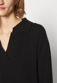 Femme portant un pull en maille côtelée noir avec un col et un décolleté en V, et une boucle d'oreille en or visible sur une oreille.