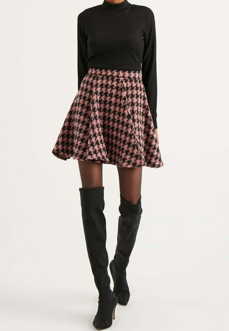 Femme portant une jupe évasée à motif pied-de-poule rose et noir, un haut à col roulé noir, des collants transparents et des bottes noires à talons hautes jusqu'aux cuisses.