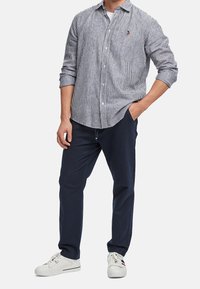 Gestreiftes Hemd in Navy und Weiß, kombiniert mit navyfarbigen Hosen und weißen Sneakers. Verfügt über ein Logo auf der Brust und hochgekrempelte Ärmel.