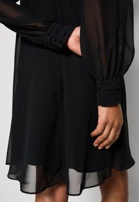 Schwarzes Kleid mit transparenten langen Ärmeln und geknöpften Manschetten. Der Saum ist leicht geschwungen und zeigt eine weiche, fließende Textur.