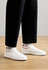 Sneaker bianchi con parte superiore in rete testurizzata, chiusura con lacci e linea di accento rossa a contrasto. Indossati con pantaloni neri a vita alta, su un pavimento in legno.