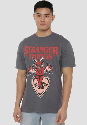 Graues Baumwoll-T-Shirt mit dem Schriftzug "Stranger Things" in Rot, ergänzt durch eine rote Teufelsgrafik, die auf einem rosa Herz sitzt, das mit "HEART CLUB" beschriftet ist.