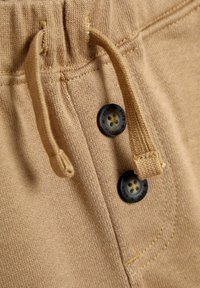 Tissu beige avec une texture douce, comportant un cordon de serrage et deux boutons ronds foncés avec un motif floral, situés près de la taille.