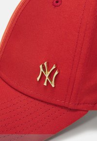 Rote Kappe aus strukturiertem Stoff mit einem goldenen Metal-Logo. Gebogener Schirm mit sichtbaren Nahtdetails und auffälligen Belüftungsösen.