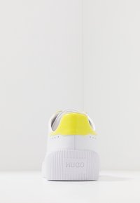 Sneaker bianca con suola scanalata, caratterizzata da un accento del tallone giallo lucido e dettagli perforati, etichettata con un discreto logo "HUGO".