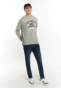 Felpa grigia con grafica "LAST FRONTIER MO UNIVERSITY", maniche lunghe, polsini a coste, abbinata a jeans blu e sneakers bianche.
