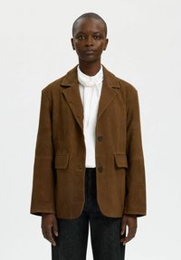 Chaqueta de ante marrón con solapa con muesca, dos bolsillos delanteros y cierre de botones. Lleva puesta sobre una camisa blanca con corbata.