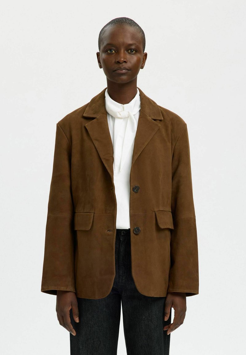 Chaqueta de ante marrón con solapa con muesca, dos bolsillos delanteros y cierre de botones. Lleva puesta sobre una camisa blanca con corbata.