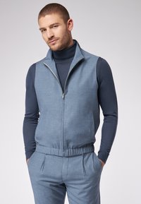 Hellblaues Reißverschluss-Weste aus strukturiertem Stoff, mit Stehkragen, Seitentaschen und lockerer Passform über einem marineblauen Rollkragenpullover.