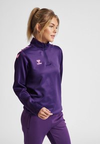 Haut de sport zippé violet avec col haut, manches longues et accents de logo rose. Fabriqué en matériau synthétique lisse adapté aux vêtements d'activités.