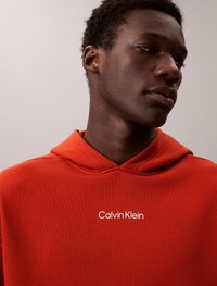 Rdeča kapuca z rebrasto teksturo, z sprednjim žepom in belim logotipom "Calvin Klein" na prsih. Kapuca z vrvico.