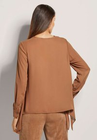 Braune Bluse mit langen Ärmeln aus weichem Material, runder Ausschnitt und seitlichen Schlitzen am Saum, getragen über passenden braunen Wildlederhosen.