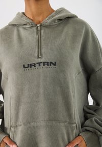 Hanorac verde măsliniu cu fermoar, echipat cu o buzunar frontal și textul 'URTRN DESIGNED IN BERLIN'. Material moale și croială lejeră.