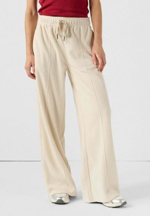 Pantaloni beige a gamba larga realizzati in tessuto morbido, con vita regolabile con cordoncino e dettagli sulle cuciture laterali. Indossati con sneakers argentate.