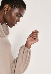 Maglione beige con collo a scialle e una consistenza morbida, con maniche arricciate, che mostra un primo piano di una mano appoggiata sul mento.
