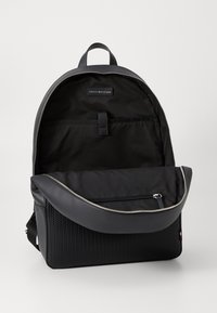 Zaino nero con parte superiore liscia, sezione inferiore testurizzata, zip argentata e dettagli del logo. Caratterizzato da un ampio interno e una tasca frontale.