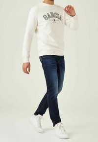 Man som bär en vit sweatshirt med texten "GARCIA", mörkblå jeans och vita sneakers, stående med ett ben över det andra.