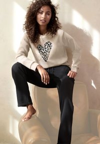 Bež dukserica s dizajnom srca u leopard uzorku, u kombinaciji s crnim, tapered trenirkama. Model sjedi na svijetloj stolici.