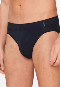 Tanga in cotone nero con una texture liscia, vita bassa e etichetta blu discreta sulla cintura. Vestibilità classica con dettagli di cucitura minimi.